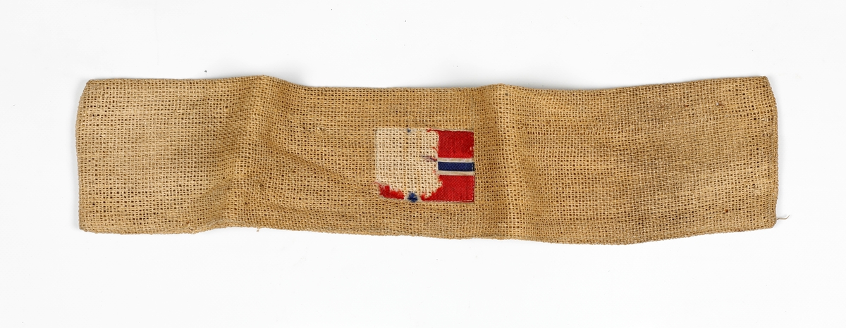 Armbind laget i vevd papir. Norske flagg sydd på. Flagget er delvis ødelagt.