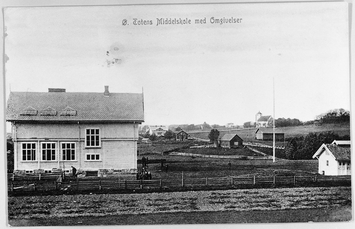 Østre Toten Middelskole med omgivelser ca. 1905. Sett fra øst mot vest. Hoff kirke i bakgrunnen.
Colorert postkort.