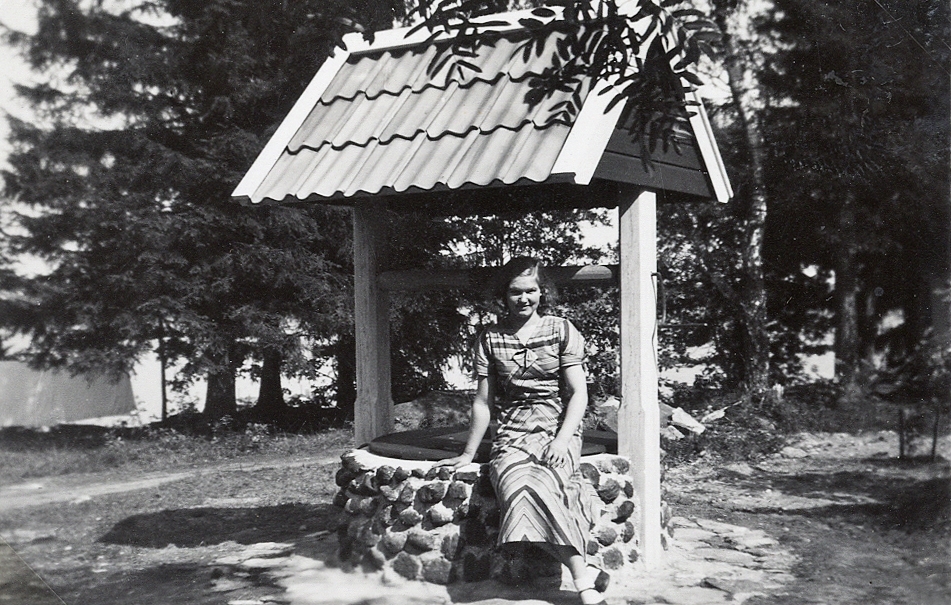 En kvinna sitter på kanten till en brunn med brunnstak.