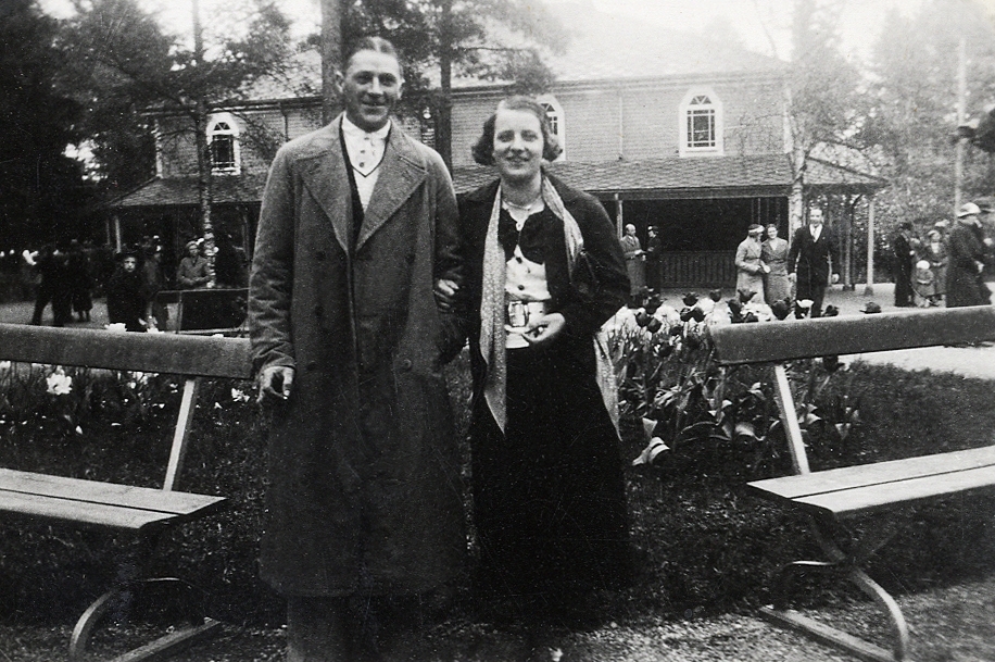 En man och en kvinna poserar för fotografen i en park. I bakgrunden syns en teaterlokal (?), restaurang (?).
