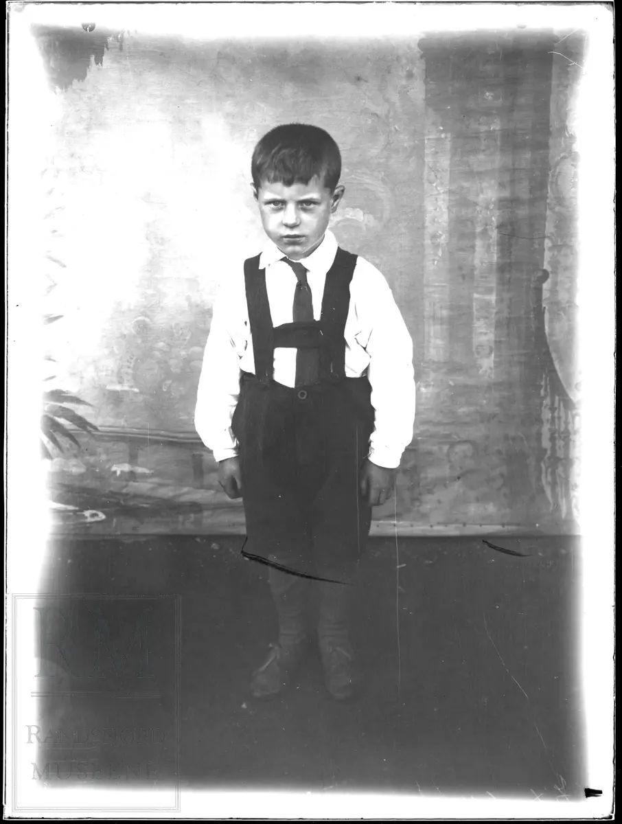 Portrett i helfigur av en liten gutt i kortbukser med sele.