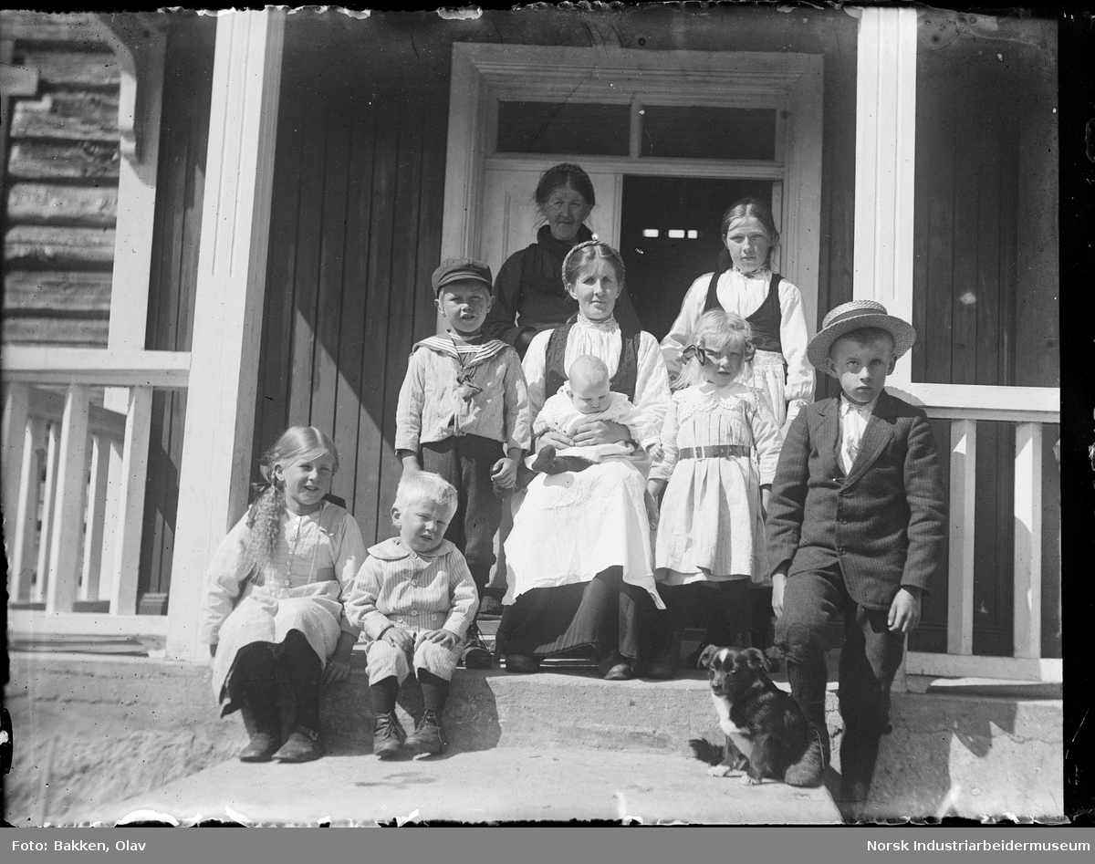 Familien Lie oppstilt utenfor inngangsdøren til huset. To voksne kvinner og syv barn. Hundevalp sittende på trappen.