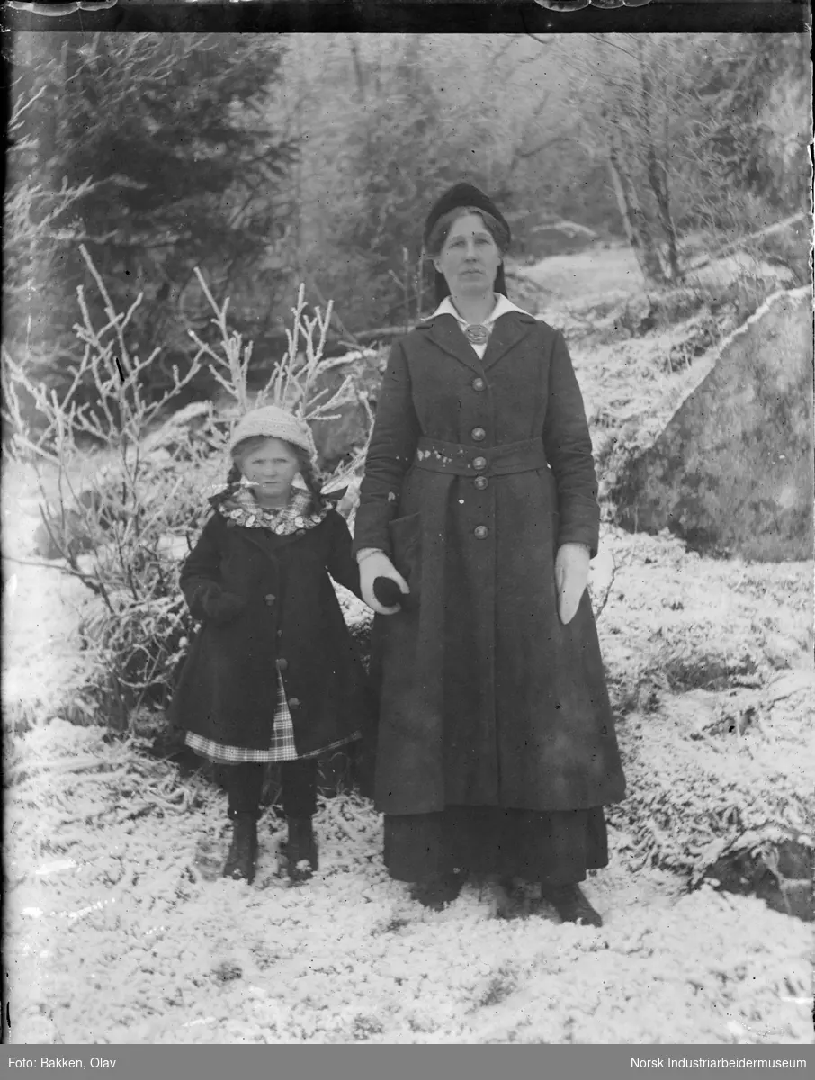 Kvinne holder jente i hånden oppstilt utendørs i snø. Begge kledd i vinterkåpe, votter og lue.