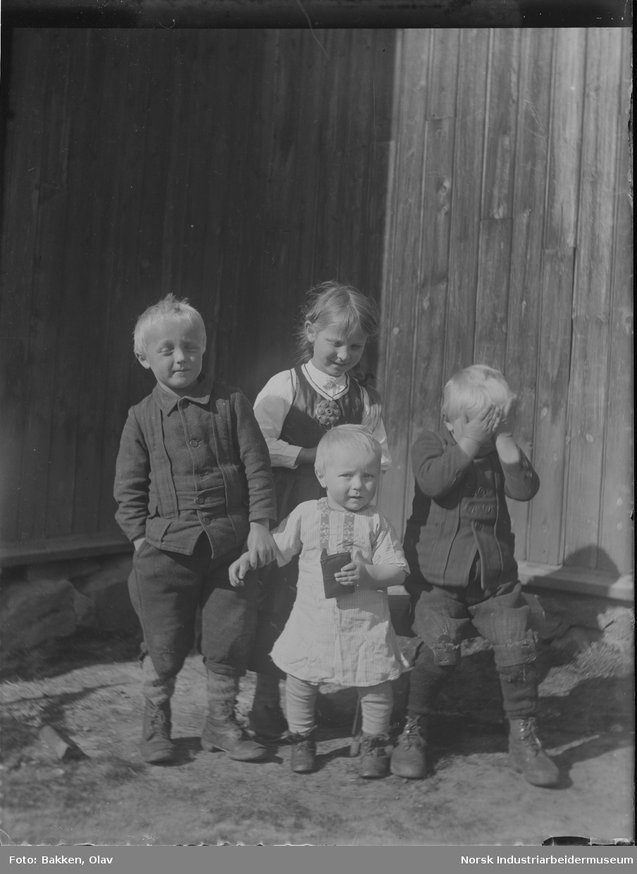 Fire barn stående foran bygg. Ett av barna holder hendene for øynene. Gutt holder mindre barn i armen.