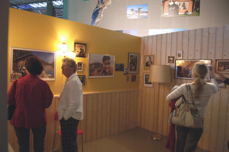 Personer ser på utstillingen. Det henger fotografier og andre bilder på en lys og en gul vegg.