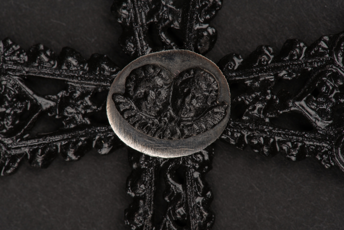Hänge till halsband i form av ett kors.
Korset är av utsirat svart gjutjärn med rombformade avslut i nygotisk stil. I mitten sitter en oval förnicklad platta med två änglahuvuden i gjutjärn.
