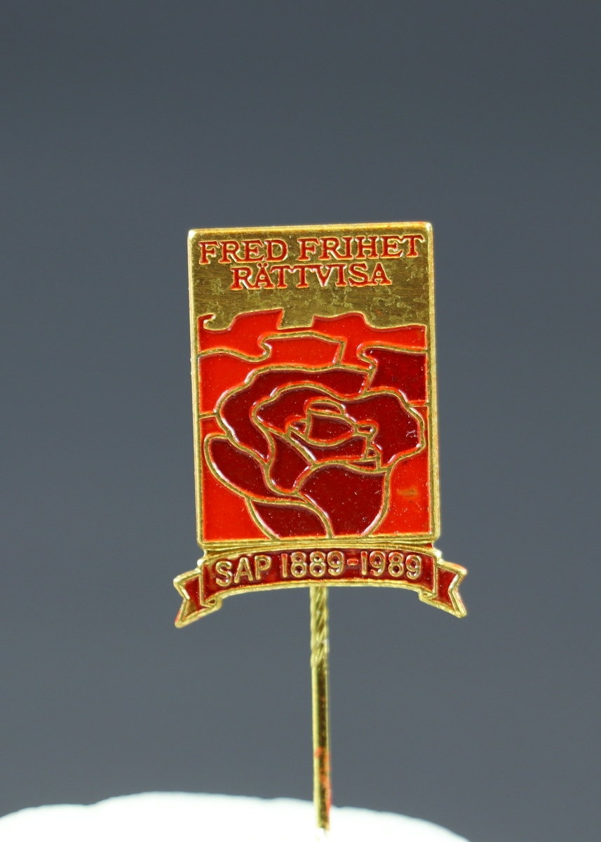 Ett fyrkantigt nålmärke i rött och gulmetall. Högst upp står det "Fred Frihet Rättvisa" under detta en röd ros och längst ner i en illustration av ett band står det "SAP 1889-1989".