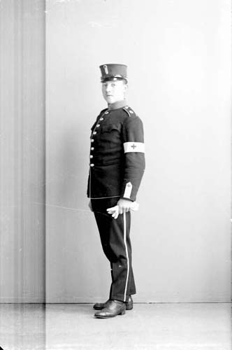 En man i militär uniform stående i 3/4-profil. Ett vit band med kors på vänster arm och vita handskar i handen.