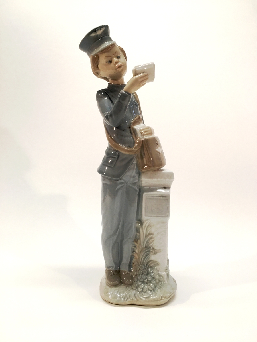 Figurin förställande en man i uniform med mössa och brevbärarväska. Han håller ett brev och står lutad mot en brevlåda.