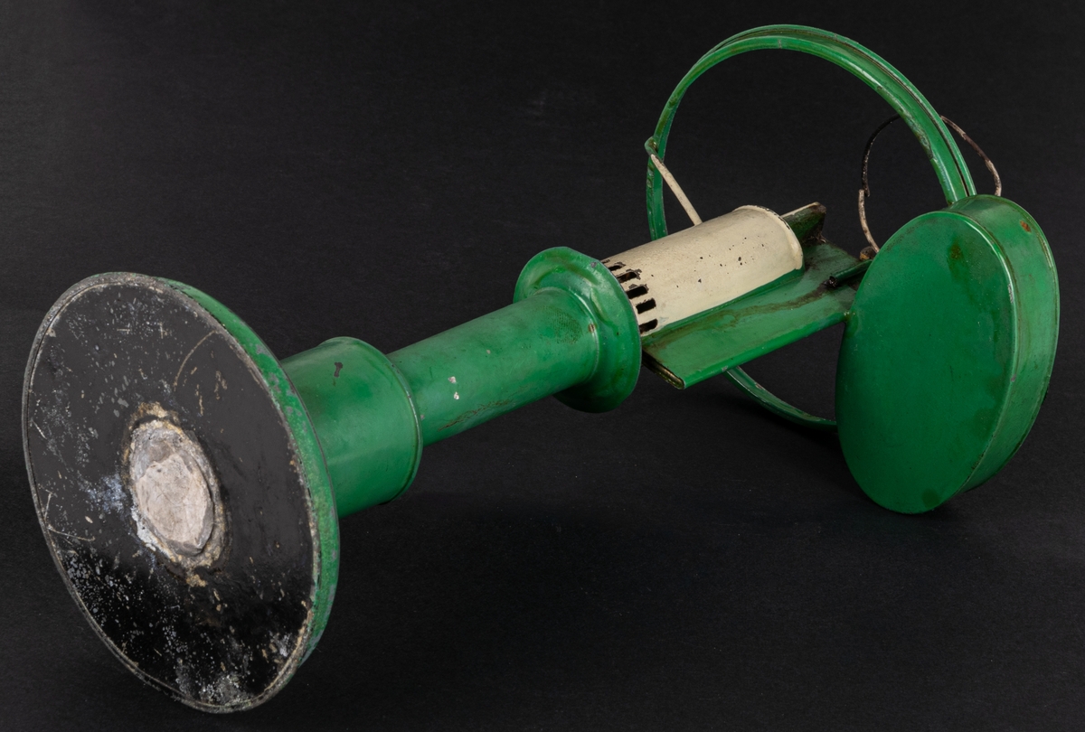 Bordslampa, liten, grön och vit.
Grönmålad plåt, på rund fot med njurformig behållare vid sidan i höjd med den lilla vita porslinskupan. Rakt, cylindriskt brännarglas.