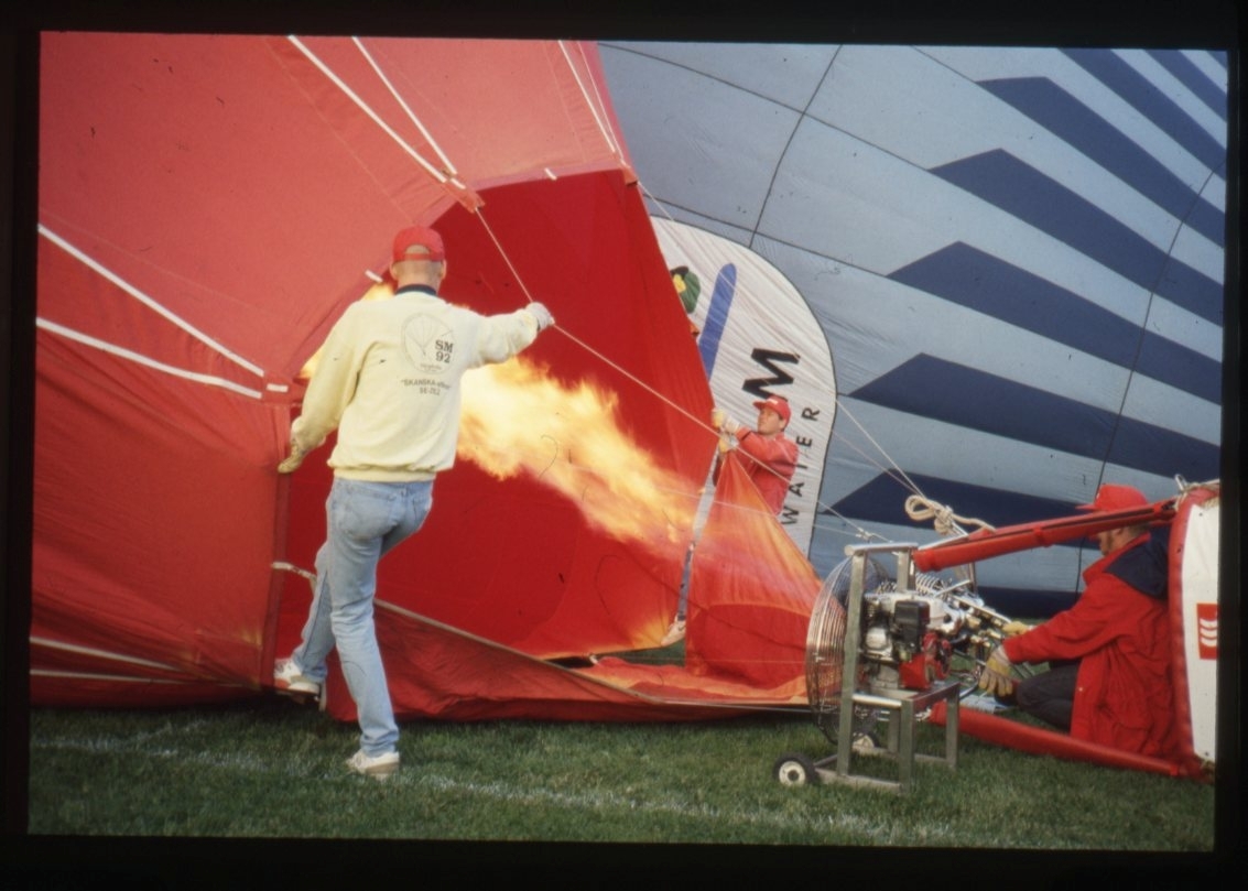 Två personer håller i ballonghöljet medan brännaren fyller ballongen med hetluft.  En eldslåga står ut från brännaren.  I bakgrunden ses ytterligare en ballong som håller på att fyllas.