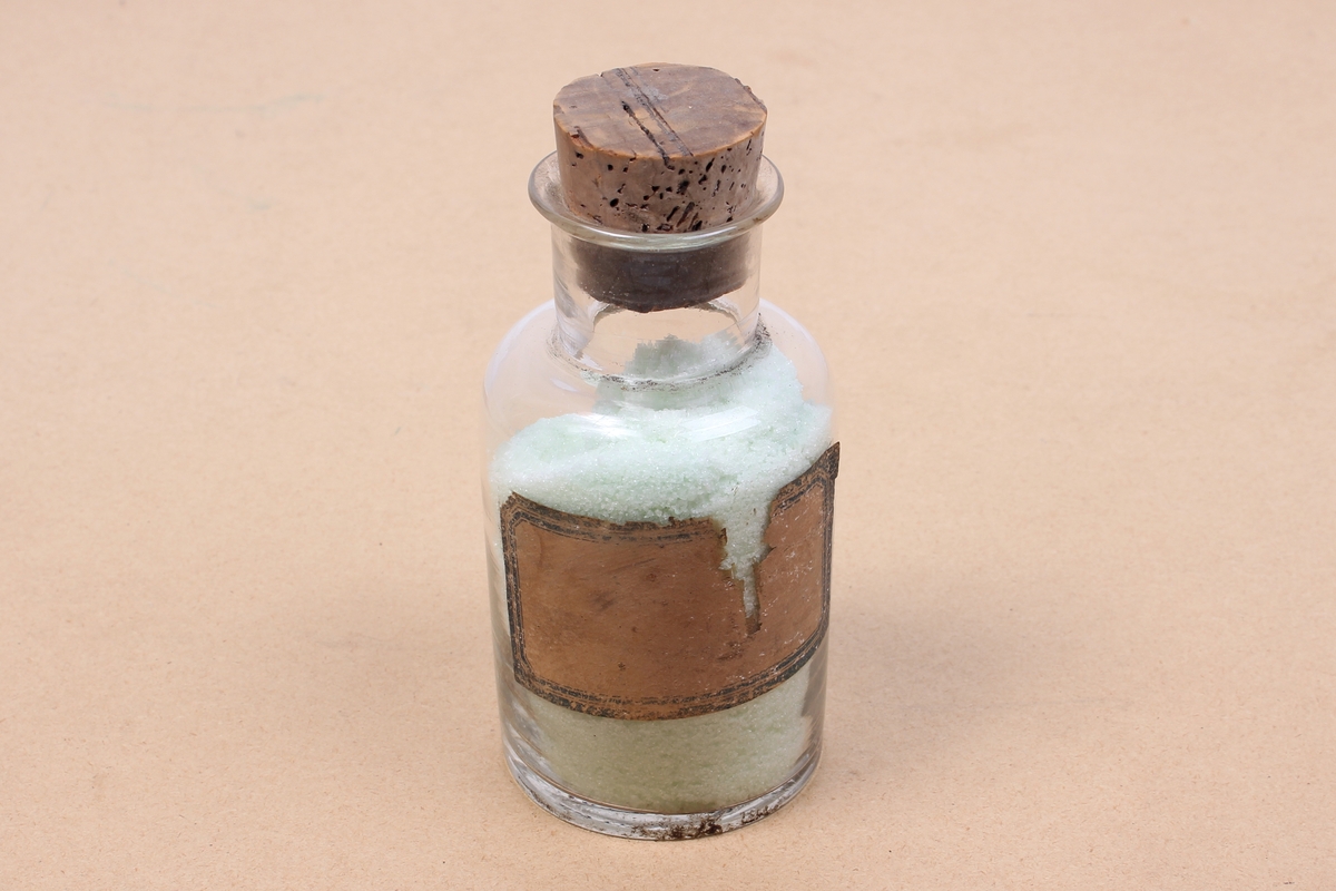 Kjemikalieflaske av klart glass, med stopper av kork. Inneholder trolig ammonium jern(II)-sulfat. Flasken har en blank etikett på siden.