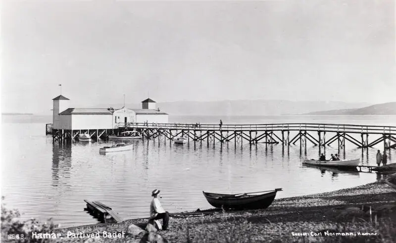 Flott badehus med to fløyer med hvert sitt lave tårn, og en lang brygge i tre som leder ut til badehuset som står på påler ute i Mjøsa.