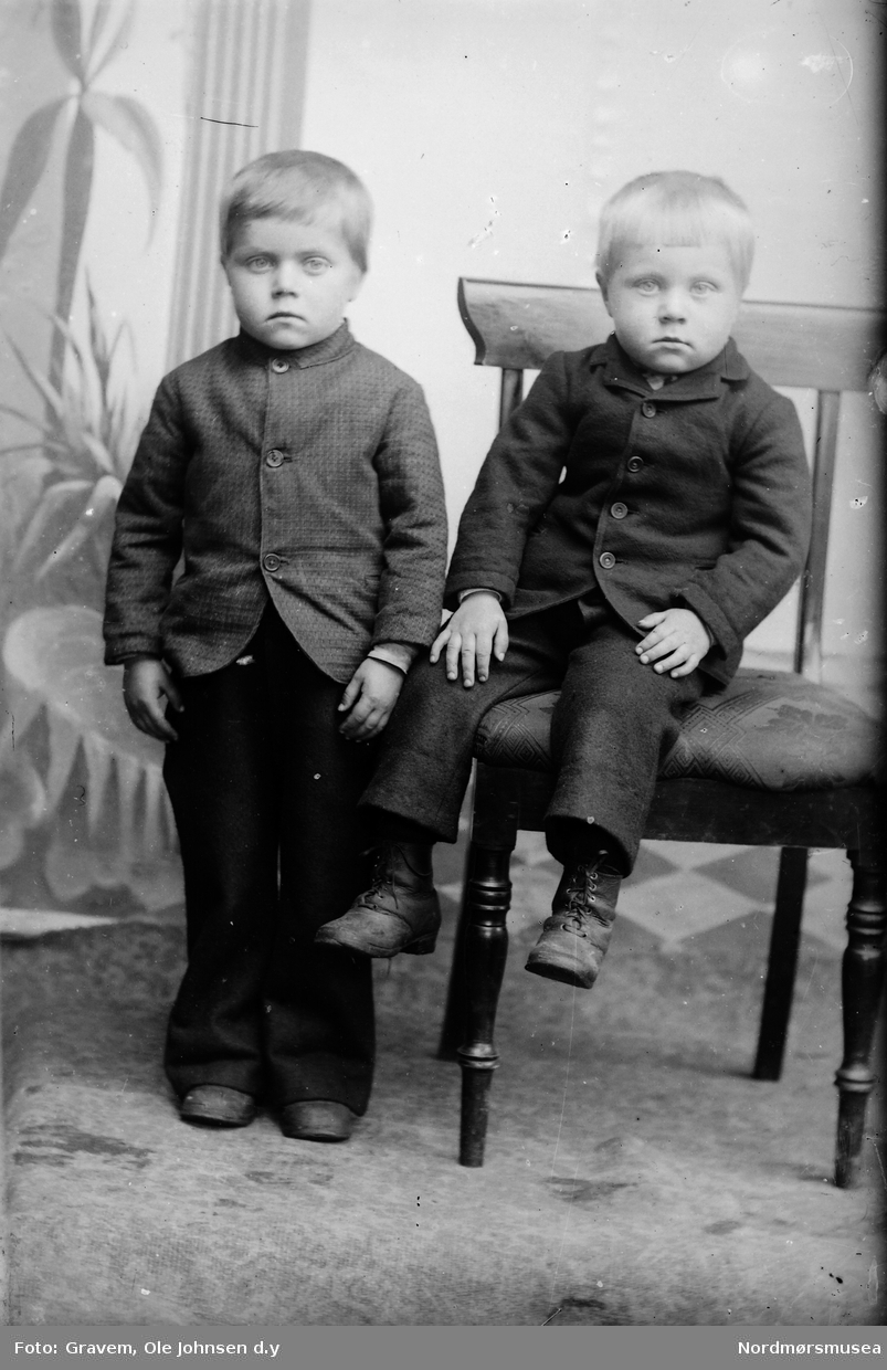 To små gutter, brødre. En sittende på stol, en stående. Atelierbakgrunn med palmer og søyler.