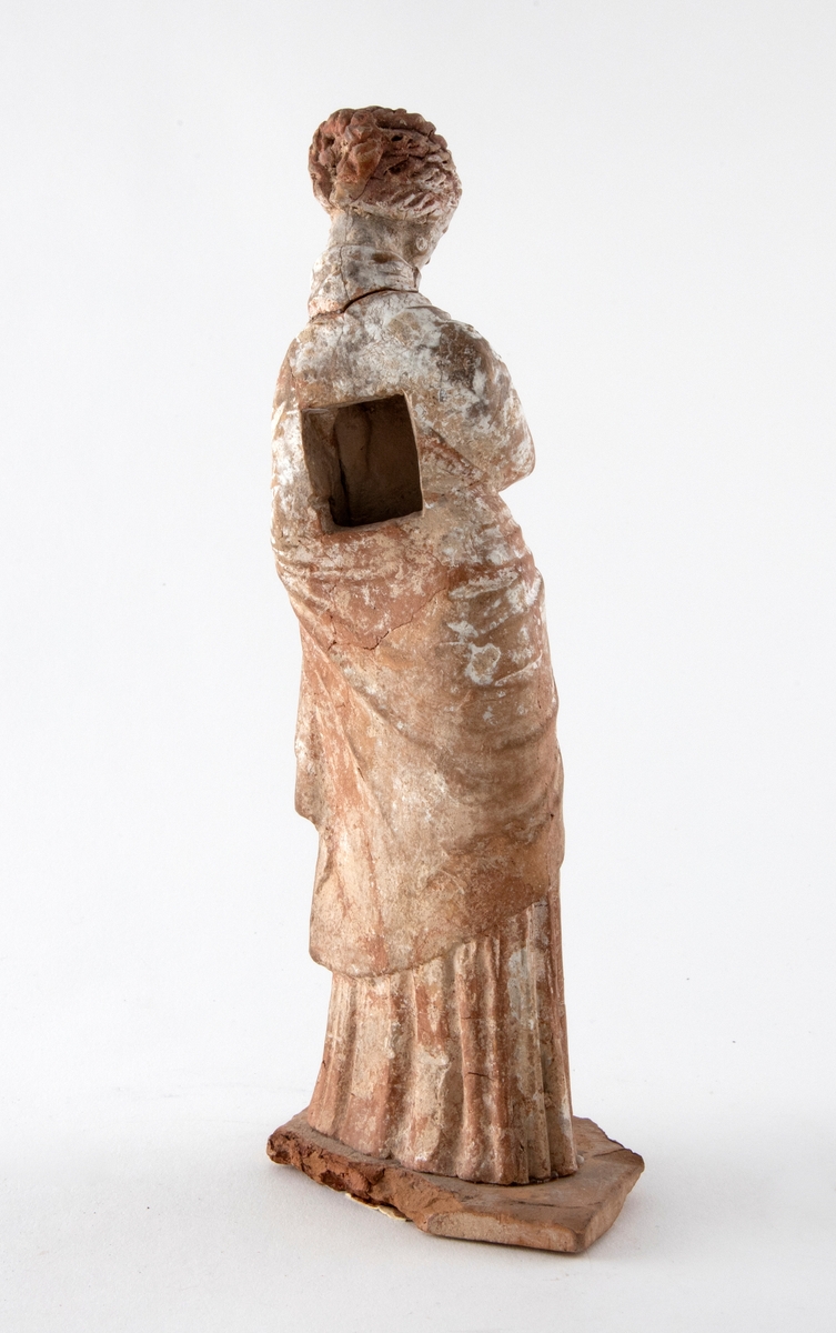 Figurin i uglasert terrakotta, dekorert med hvit begitning. Figurinen er formet som en stående kvinne, iført himation. Rektangulær åpning i ryggen, og innsiden er hul. Figuren står på en fotplate av terrakotta.