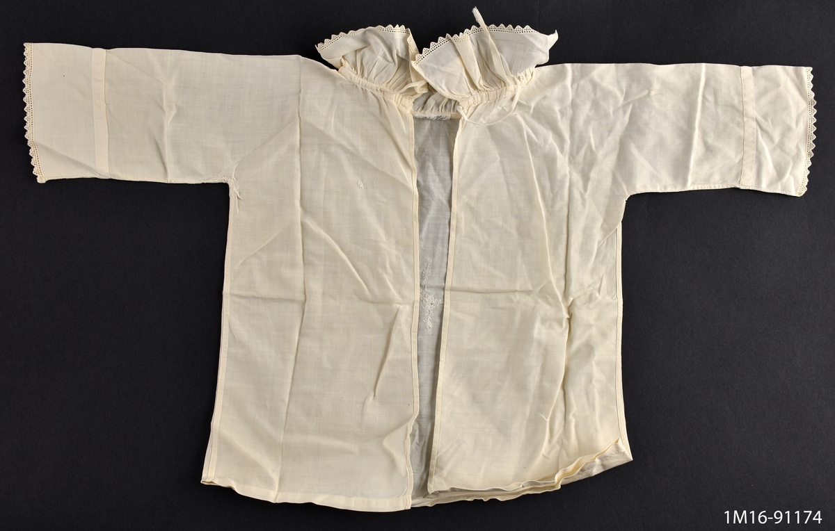 Skjorta till baby, vit, öppen bak, knytes ihop med vitt bomullsband vid kragen, helskuren med långa ärmar, dessa och kragen prydda med smal uddkant. Lagad fram.