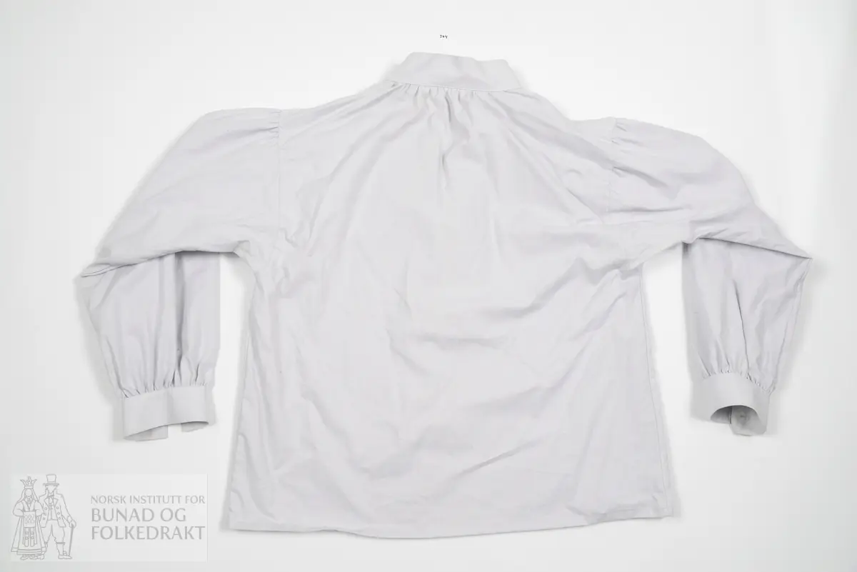 Skjorte i grå bomullslerret til kvinne. Moderert primærsnitt. Bol med skuldersøm og lett buede ermesømmer. Ekstra vidde rynket til formklippet halskrage, splitt foran med vrangsydd belegg. Ermene er klipt i lett bue nærmest bolen. Rynket ermetopp, rynket til ermekrage. Maskinsydde doble knapphull. Skjorta er maskinsydd, konfeksjonssøm på størrelse.
