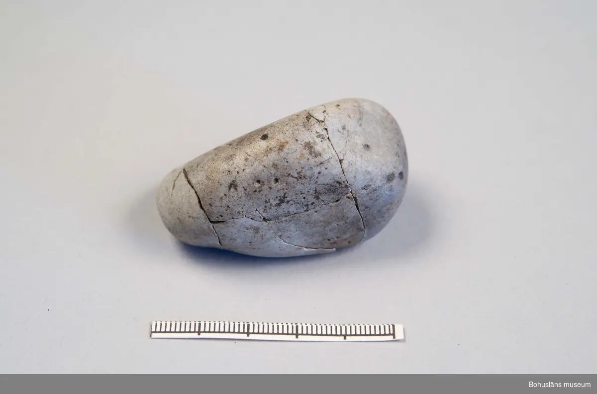 Slät oval grå sten med en i det närmaste polerad yta. Funnen i en grav och delad i fyra delar som låg på olika platser i brandlagret. Numera är den sammanfogad till ett föremål.