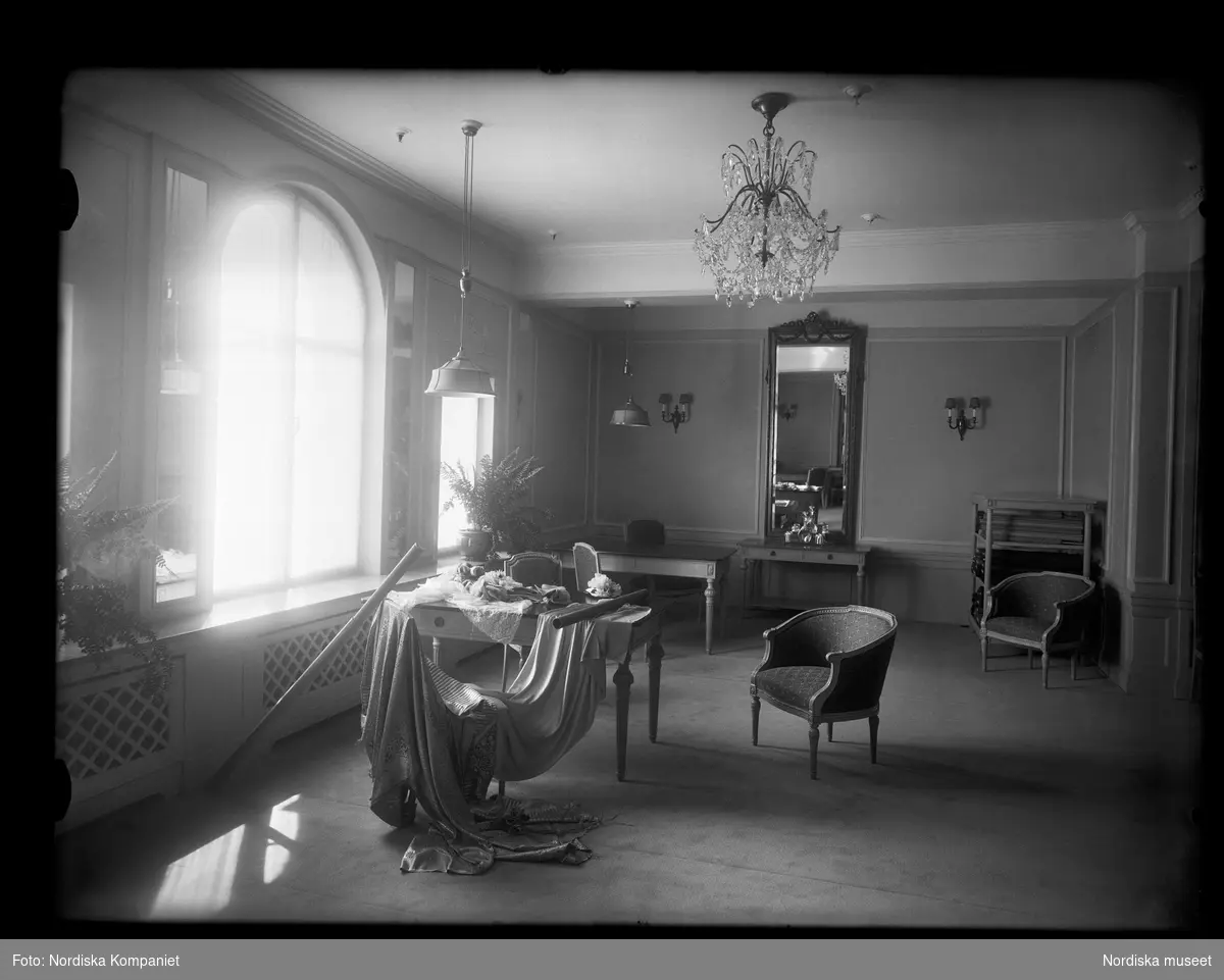 Interiör från Franska damskrädderiet på Nordiska Kompaniet, våningsplan fem. Rummet är elegant möblerat  med stilmöbler och en stor spegel med förgylld ram. Till vänster i bild ett stort fönster med välvd överdel, där solen strålar in. På ett bord ligger provbitar av textil och ett tygsjok ligger draperat över en stol. 1925-1929.