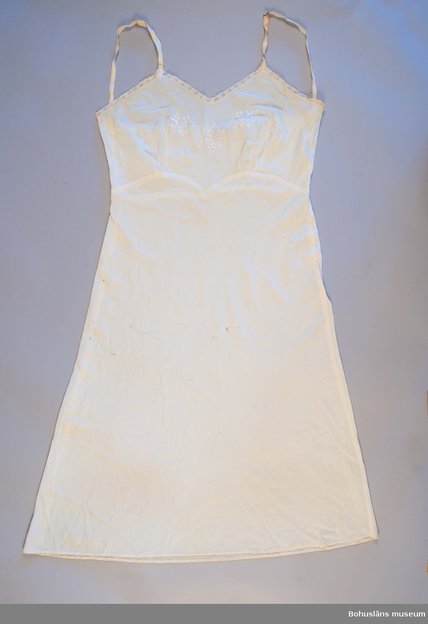 Underkläning av rayon med maskinbroderi i vitt. Modell avskuren under bysten med formsydda kupor.
Tillverkad i USA.