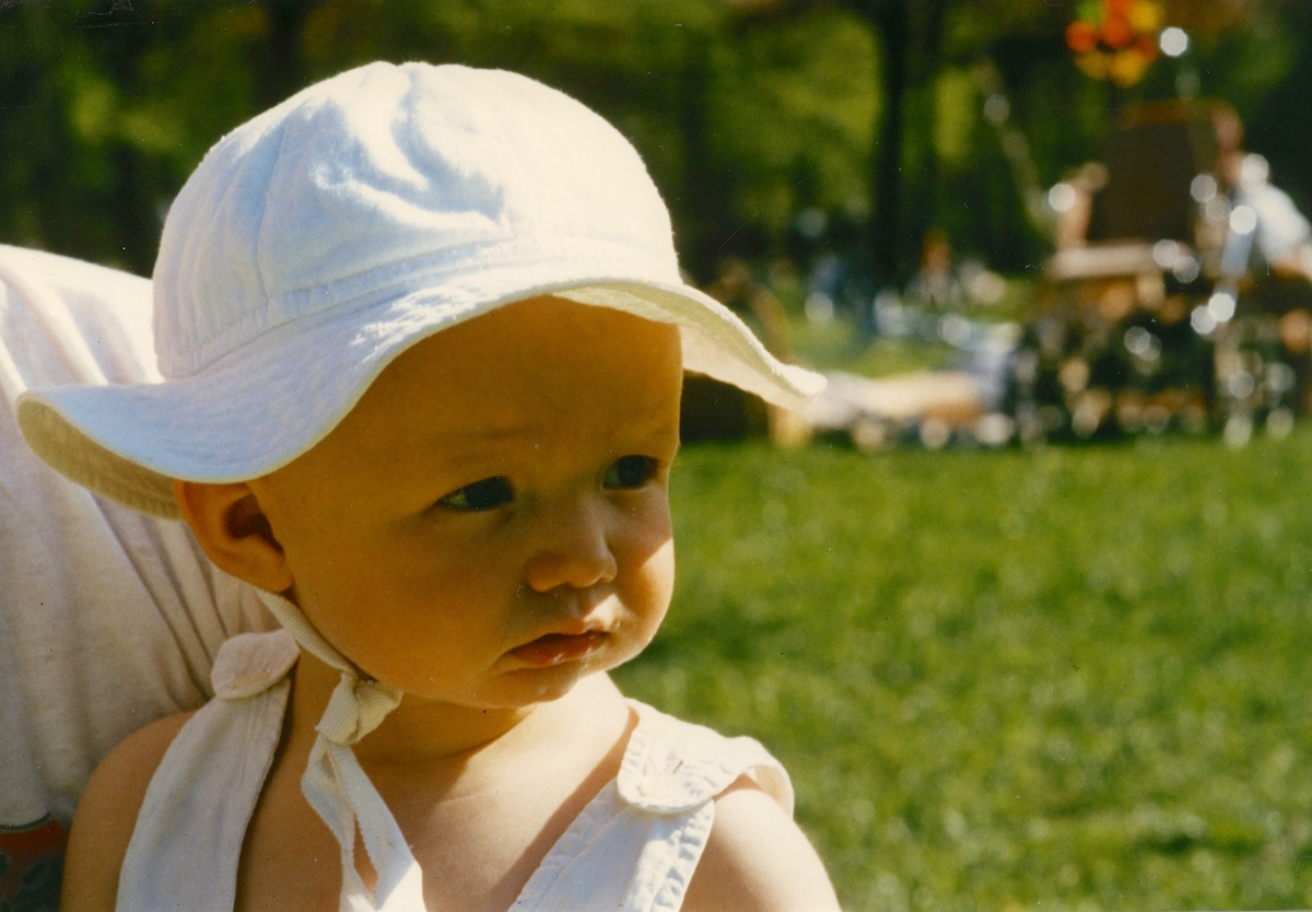 Emelie Ekman (född 1987), iklädd solhatt, sitter i någons knä i trädgården, Vommedal Östergård "Petters" på Streteredsvägen 43, år 1988.