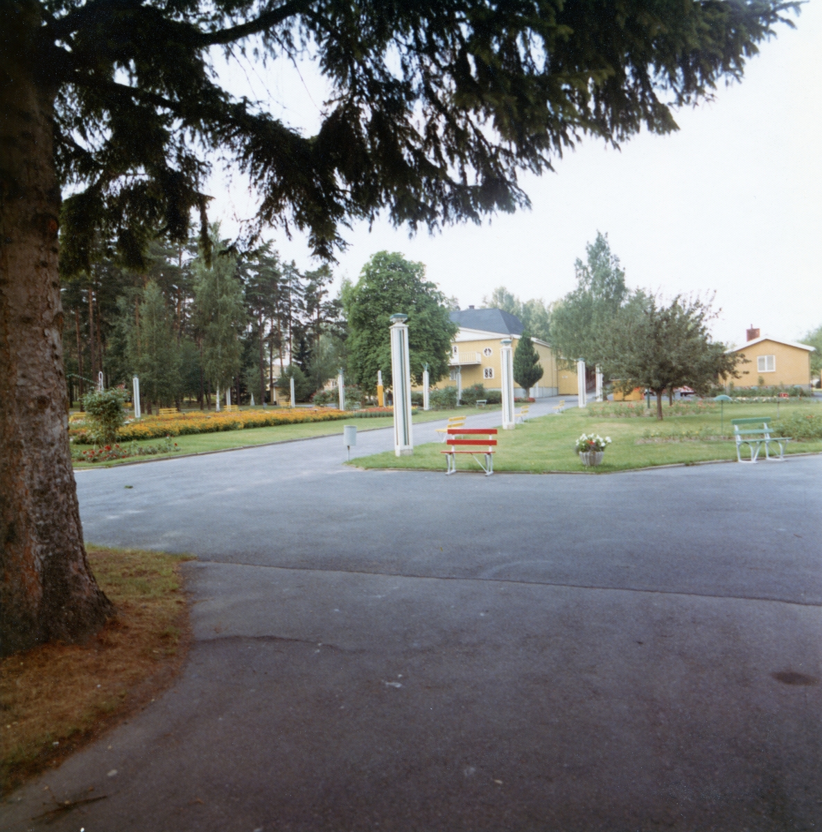 Bild från parken i Folkets Park. I bakgrunden är teatern. Gräsmattor, rabatter, parkbänkar, vita pelare och asfalterade gångar.