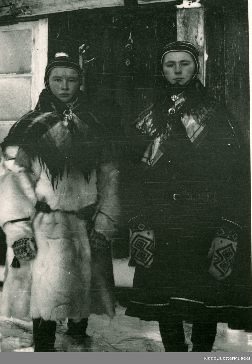 Guokte sámi nieidda nuppis gábabeaska olgun.
To kvinner utenfor et hus.