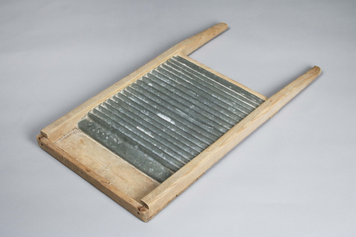 Tvättbräde. Ram av ofärgat trä, med själva tvättbrädan av räfflad, galvaniserad plåt. Övre listen med träbortfall.

Funktion: 
En tvättbräda är en räfflad skiva av metall, trä eller glas i en träram, med vilken man tvättar kläder. Detta görs genom att gnugga tyget med hjälp av en borste fram och tillbaka på tvättbrädan som hålles i en balja med vatten. (Wikipedia)
