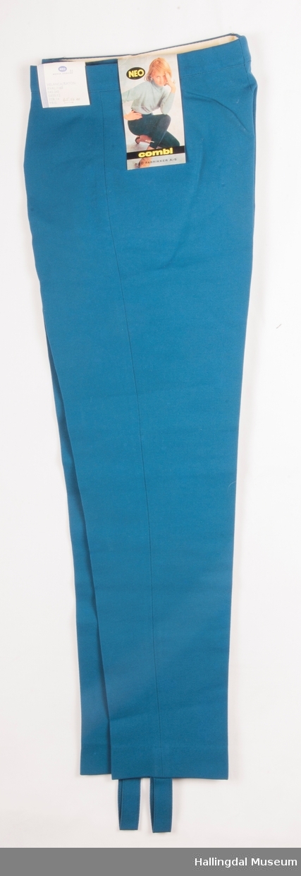 Tyrkis strekkbukse i Helanca/Rayon med glidelås i gylfen.  Fotstroppen er i samme tøy som buksa.  En lomme på høyre side framme med nylonsfor.