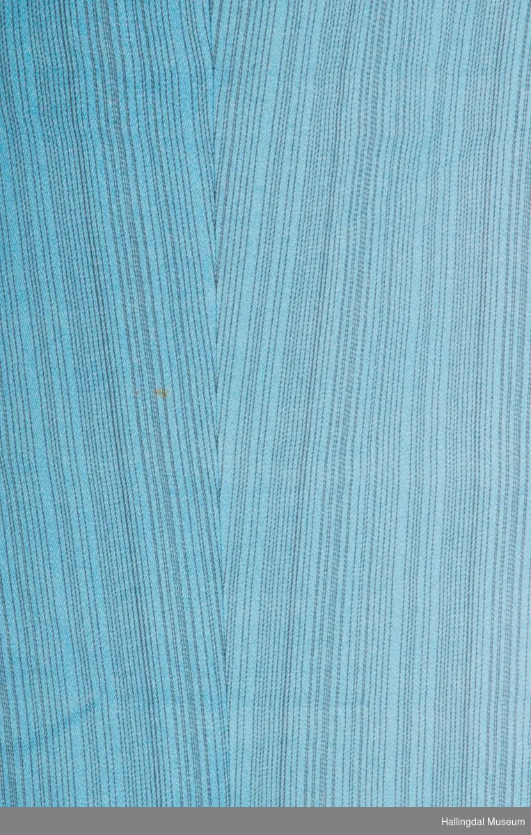 Grønn syntetisk (44% Helanca og 56% Rayon) bukse med svarte smale striper. En liten lomme framme på høyre side med klaff og knapp til lukking.  Metallglidelås i venstre side.  Spensel med 2 knapphull i linningen bak som strammes i knapper på linningen framme.  Smal i foten.