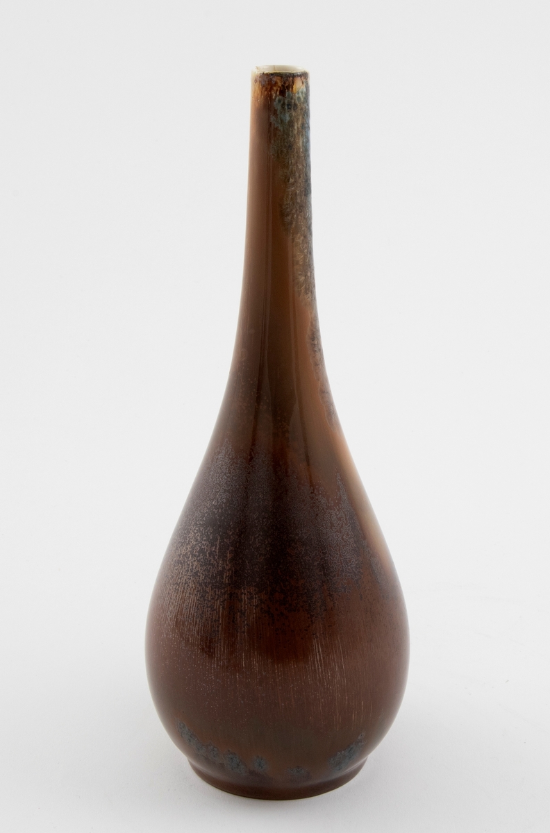 Vase i støpt porselen dekorert med krystallglasur. Dråpeformet korpus med en metallisk glans som glir over i en lang tynn hals, hvor det er små okerfargede krystalliseringer på den lange halsen.