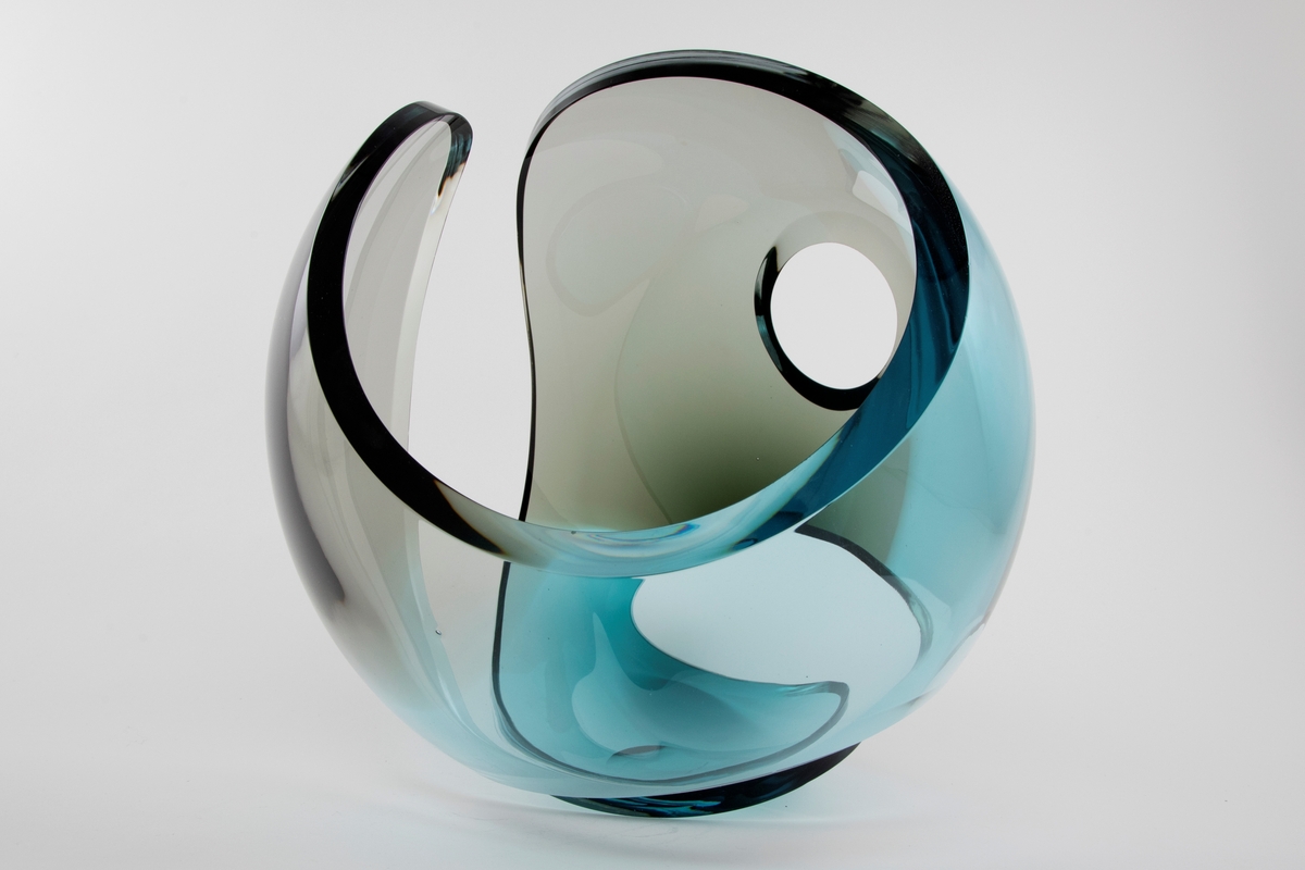 Ovalformet gjennombrutt skulptur i klart og farget glass. De blå og grå partiene overlapper delvis med hverandre, slik at det oppstår en optisk illusjon.