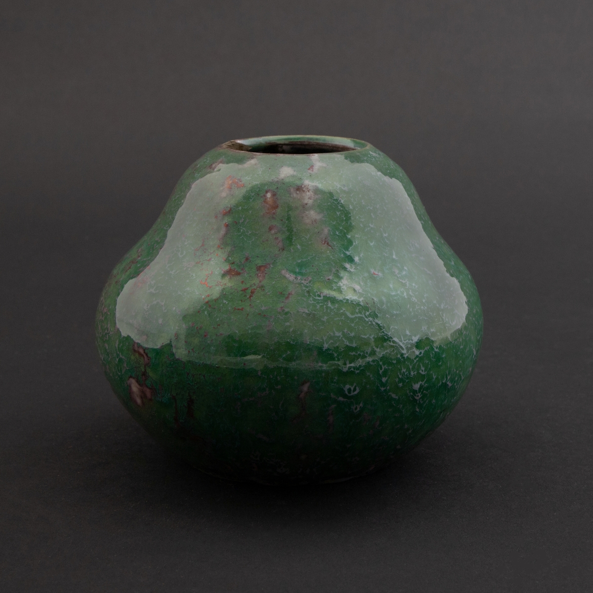 Flattrykket kalebasslignende vase med liten sirkulær åpning. Grønnfarget overflate med tidvise innslag av en mørkerød metallisk lusterglasur.