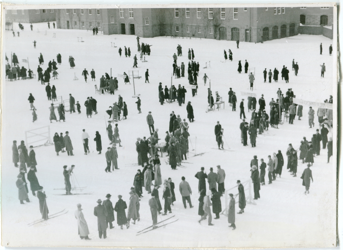 Besöksdag I 4 1900-tal. Garnisonsbyggnader i vinterlandskap med skidåkare.