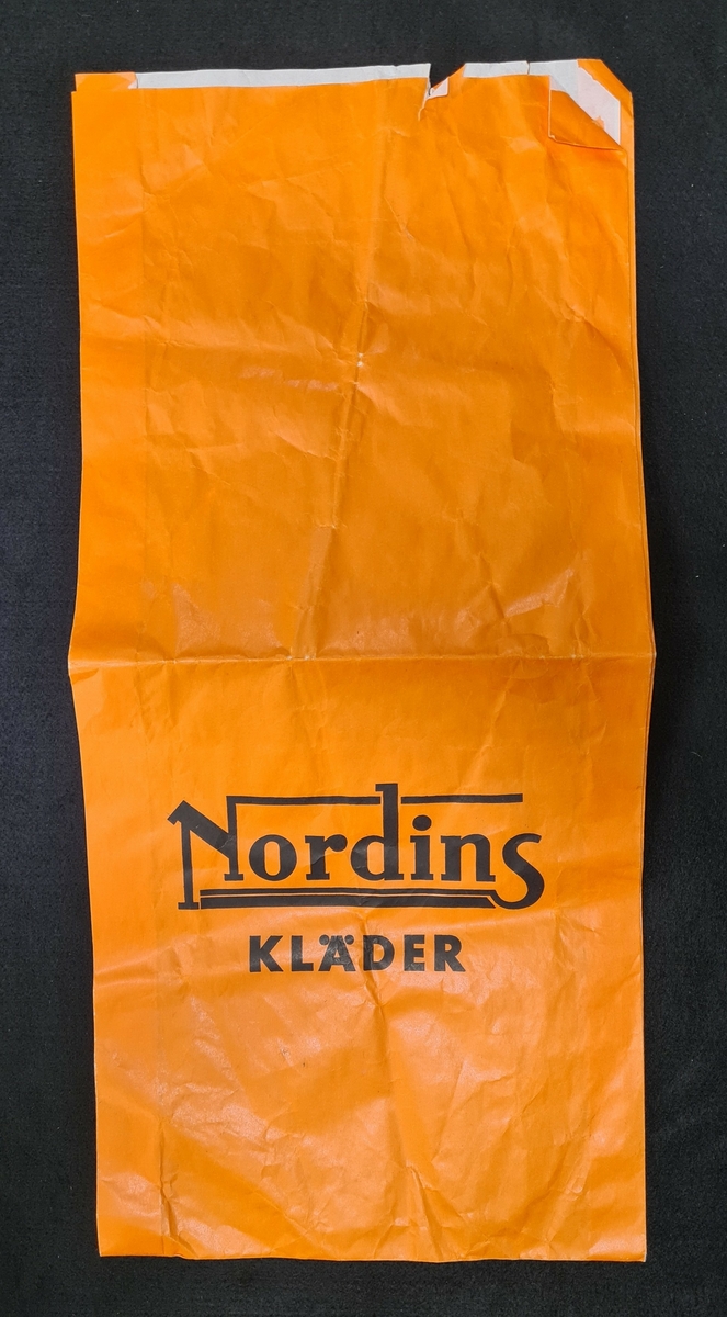 Papperspåse, orange, med logga från Nordins kläder. 

På påsen står:
Nordins kläder