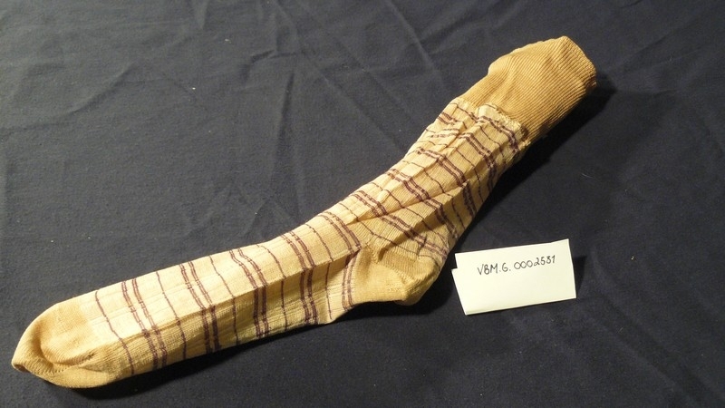 Strømpe strikket i bomull, beige med lilla striper i hele strømpen, bortsett fra tåen som er ensfarget beige. Oppe i leggen er det sydd fast en vrangbord, 9,6 cm, med foldesting.