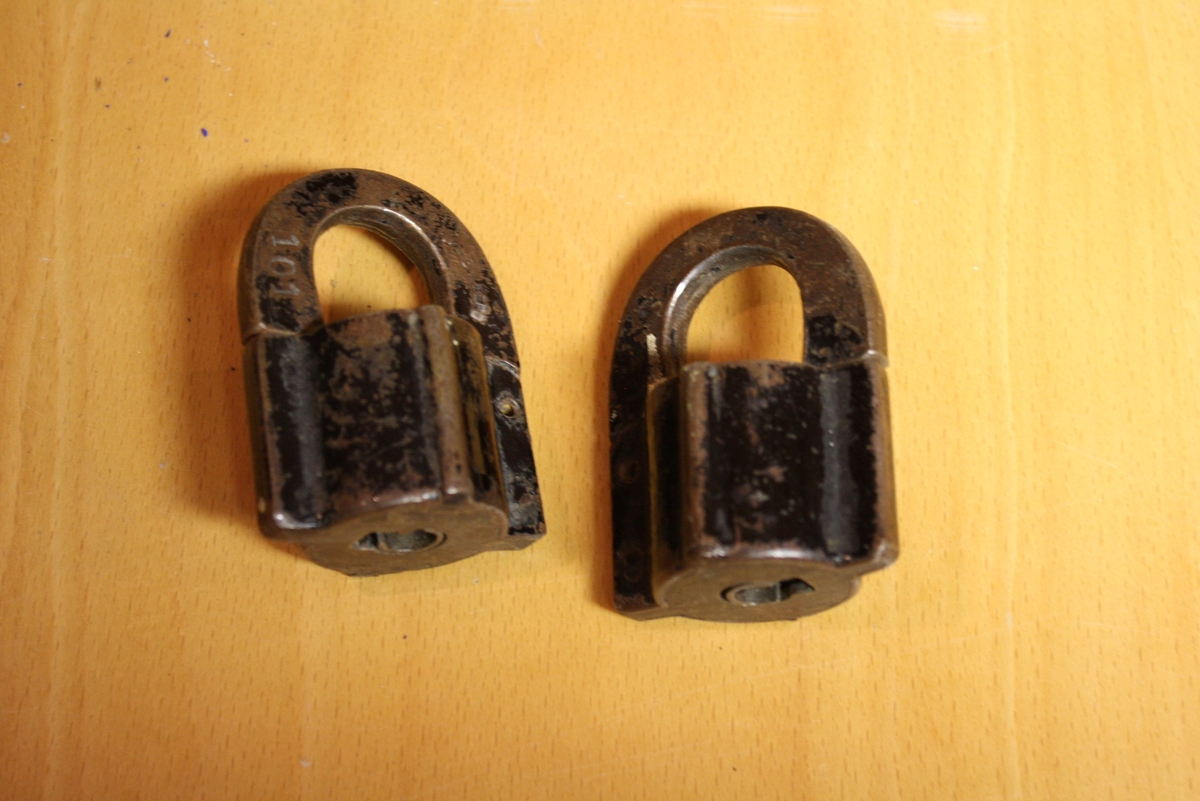 Två små hänglås i metall med svartlackerad yta. Lås på undersidan, nycklar saknas. Färgen är ganska avskavd.