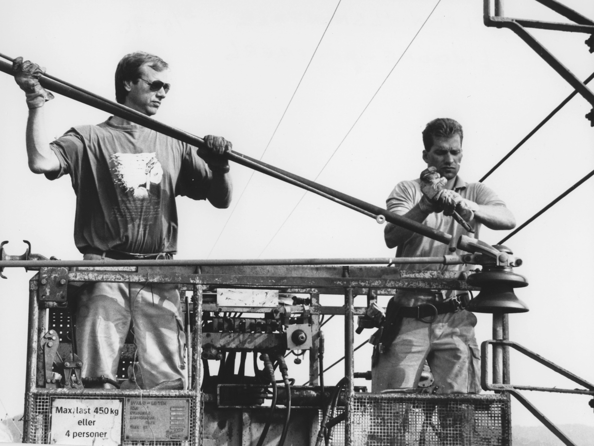 Närbild av två okända män som monterar kontaktledningar stående i lyftkorgar fastsatta i tåg. De befinner sig över järnvägsspåret i Anneberg, Lindome, hösten 1990-08-31. Relaterat motiv: A3052.