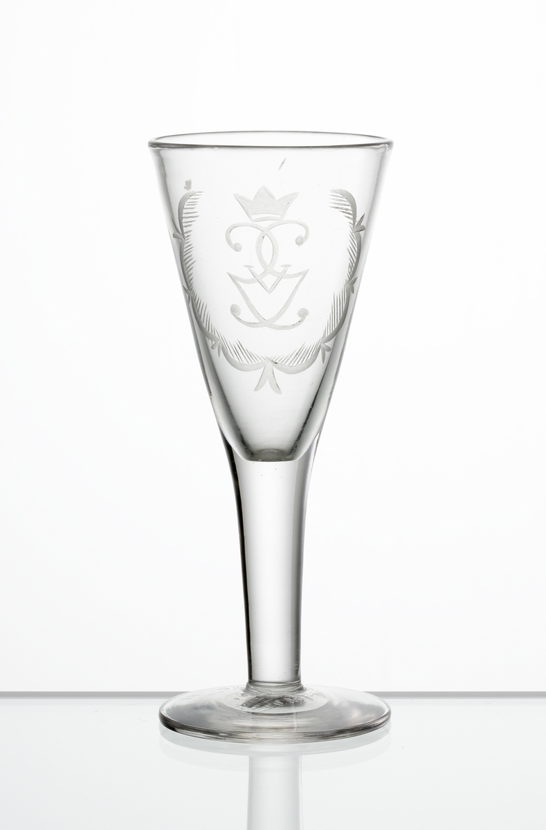 Design: Edward Hald.
Brännvinsglas. Konisk kupa med graverat monogram under krona (Gustaf V) omgärdat av stiliserad girland. 

Gustaf V (1858-1950), kung av Sverige 1907-1950.