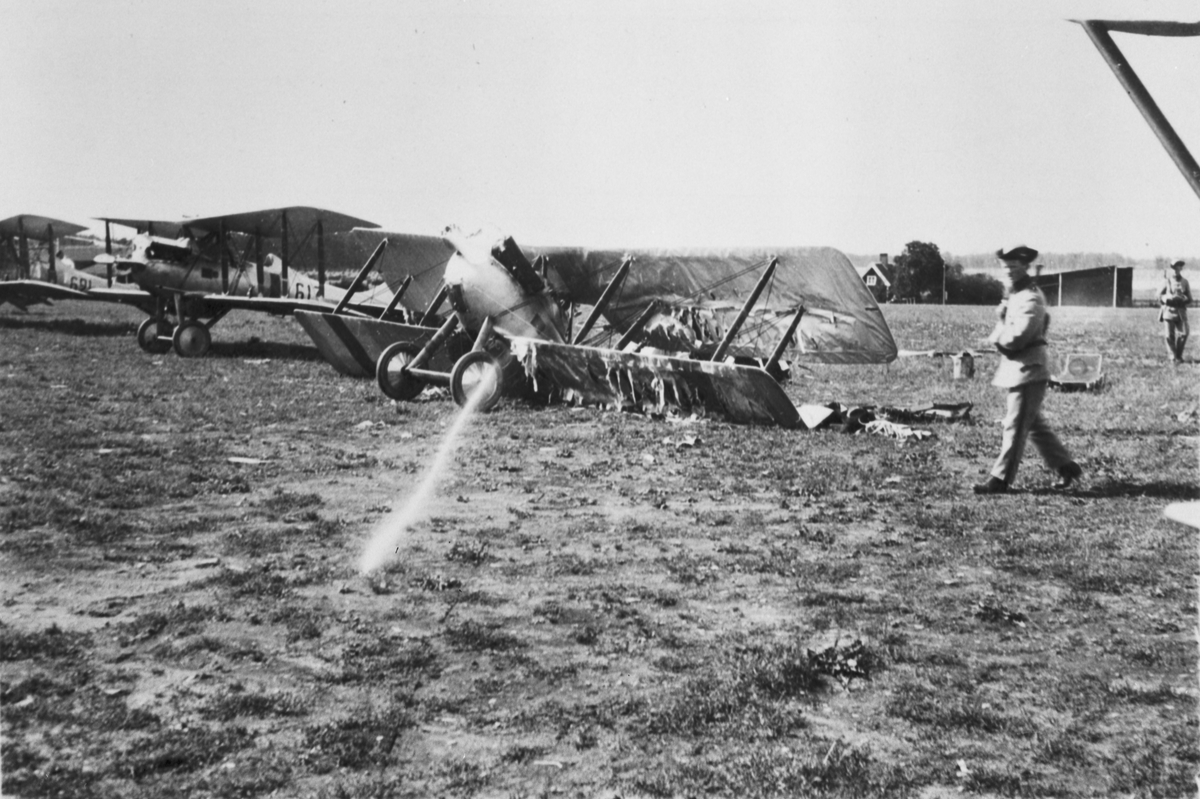 Flygplan J 2 nummer 67 står på Flygkompaniets flygfält på Malmen efter explosion vid luftpåfyllning den 15 augusti 1926. Två militärer bredvid flygplanet. I bakgrunden syns flyplan J 2 nummer 617 och 621.
