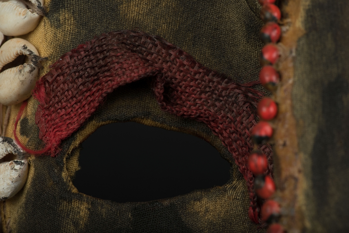 Rektangulär mask tillverkad av en dekorerad överdel av en plastdunk.
Den avskurna dunken är klädd med textil, troligen ett nylontyg, som är delvis svartmålat. Två utskurna hål bildar ögonen på var sida om handtaget som utgör näsan. Dunköppningen därunder blir en mun. Över ögonen sitter pålimmade vävda röda band i plast som ögonbryn. 
Runt ansiktet sitter en rad med kaurissnäckor och runt handtaget/näsan samt på en rad mellan mun och näsa finns fastlimmade röda frön eller bönor.
Snäckorna är fästade med tråd och på något ställe med metalltråd. Bönorna är trädda på ett snöre, sedan limmade.