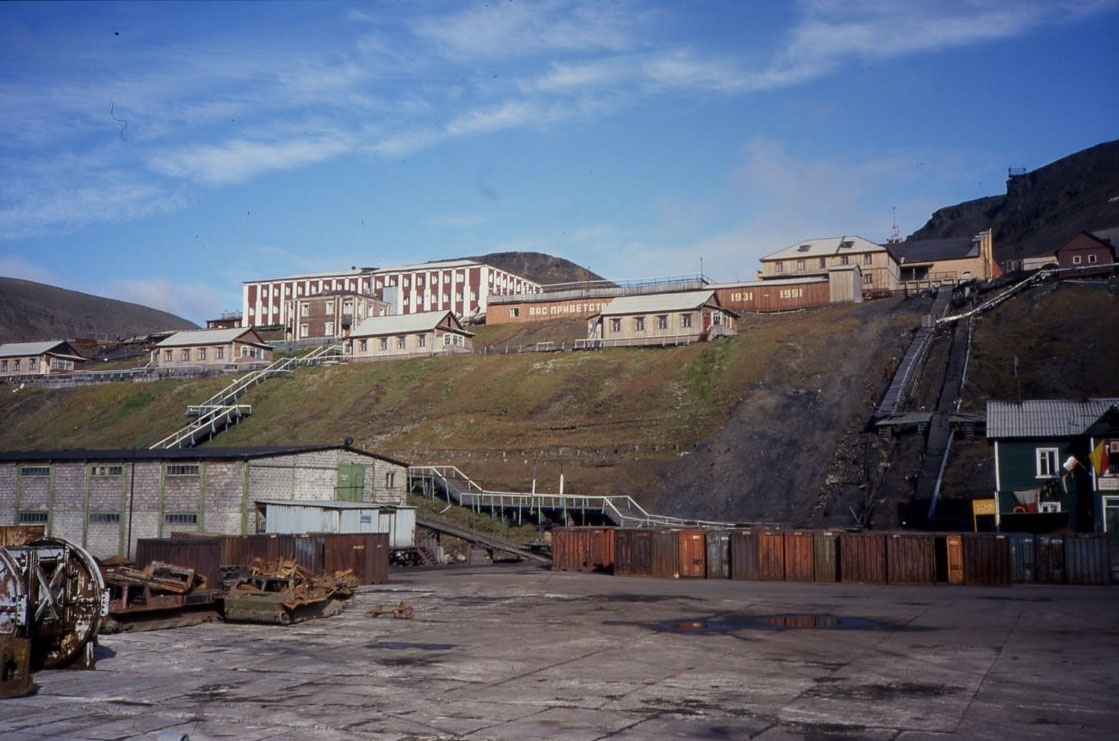 Det ryska gruvsamhället Barentsburg fotograferat från kajen. Containrar och lite järnskrot. Större och mindre byggnader ovanför en slänt.