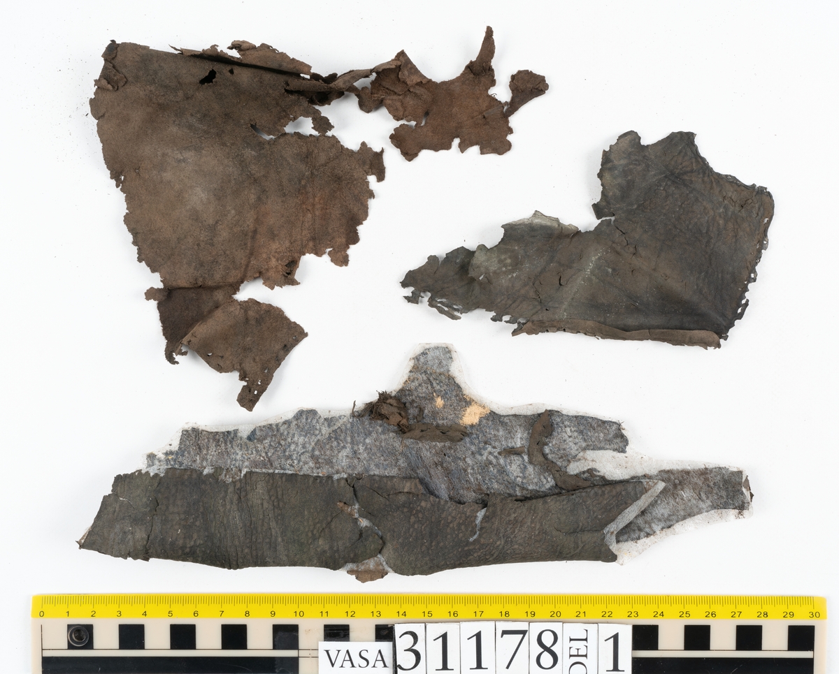 Runt 400 delar och fragment från skor i form av sulor, inlägg, ovanläder, klack, sidostycken, bakkappor, träpliggar, tråd mm. Troligtvis rör det sig om delar och fragment från flera skor och/eller stövlar. Delar av lädret har förstärkts med lim av konserveringsenheten. Merparten av lädret är spaltat samt har hål efter träpligg eller stygn.