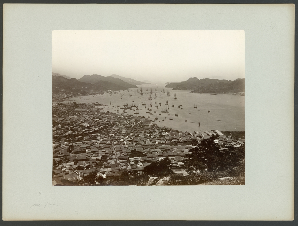 Bilden föreställer en panorama från Nagasaki. Den är tagen från en förhöjd ståndpunkt på kullarna ovan på staden. I förgrunden syns staden låga träbebyggelse och i bakgrunden bukten och hamnen fullt med båtar och skepp.