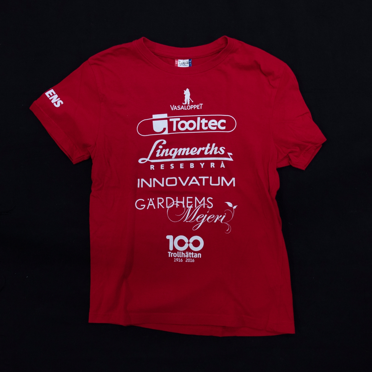 T-tröja, eller T-shirt, röd. Använd i skola i gymnastiken. 
Reklam är tryckt på tröjan från flera sponsorer.