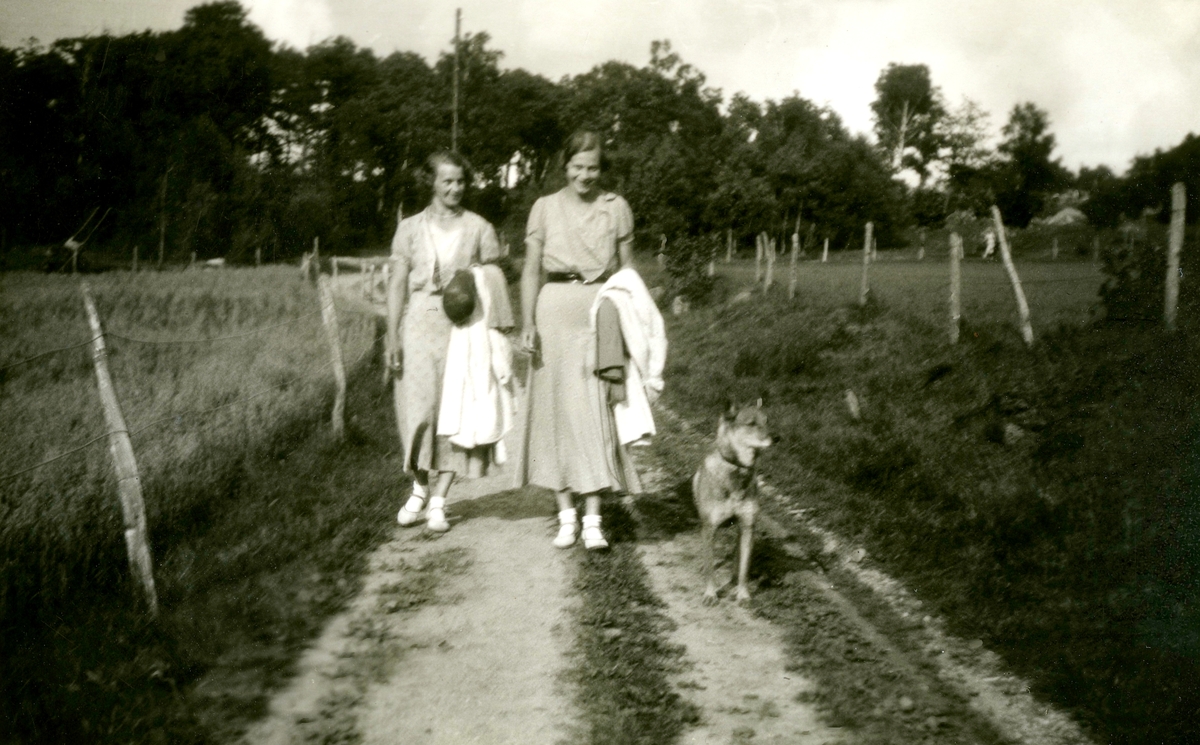 Systrarna Astrid (1907 - 1994, gift Jägerström, Råda) och Ingeborg Gustafsson (1901 - 1987, gift Johansson) promenerar på en landsväg tillsammans med en hund i koppel, Kållered Stom "Nygård" okänt årtal.