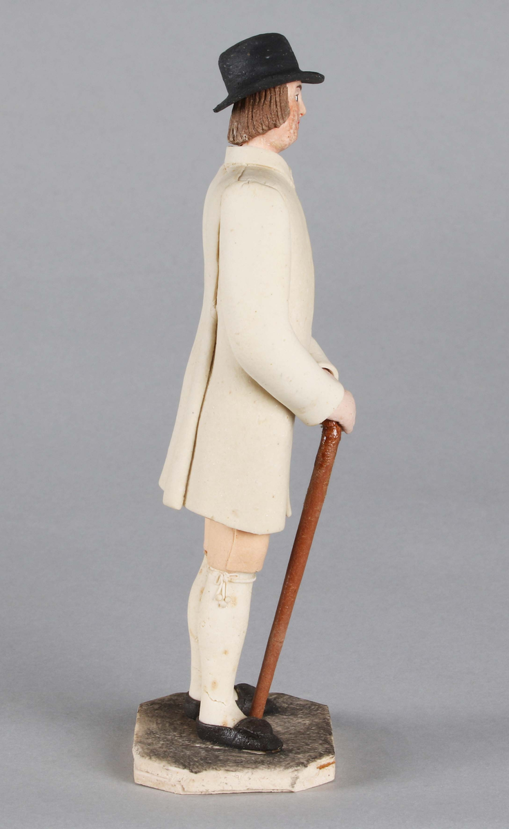Dragantarbete. Mansfigur.
Allmogedräkt. Stående mansfigur iförd svart hatt med platt kulle och rakt brätte, ej så brett. Rakt kortklippt hår. Halsduk i halsen. Ljus 
ytterrock med långa ärmar, i vars mittparti synes randig väst, röd och svart. Knäkort, ljus, aprikosfärgad byxa, med vita tofsbollar vid knäet. Vita strumpor. Svarta skor. Brun promenadkäpp. Står på grått underlag bestående av åttkantig platta.

Tillverkad av Hilda Boström (1822-1903), sockerbagare, Borås.

Litt: Se Årsbok 1963, sidan 44.

Funktion: Prydnadsfigur