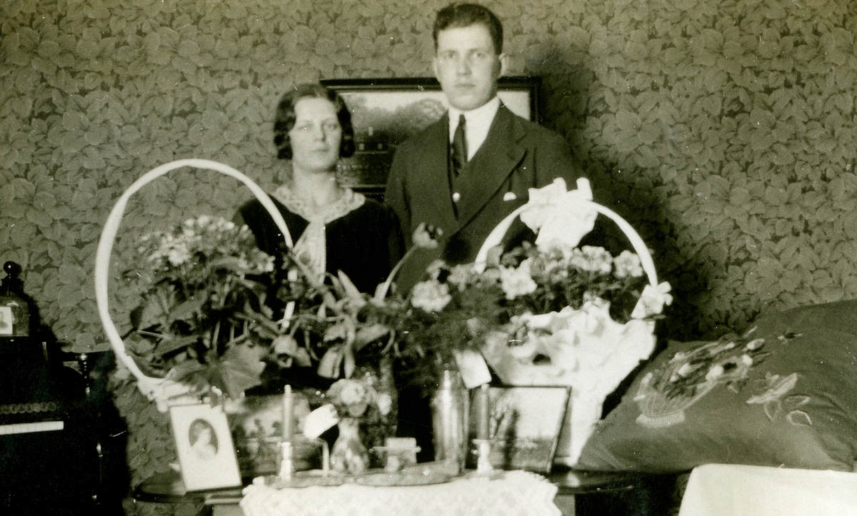 Uppvaktning för en man och en kvinna som står bakom ett bord fullt med blommor, Kållered Stom "Nygård" okänt årtal. En mönstrad tapet ses bakom paret.