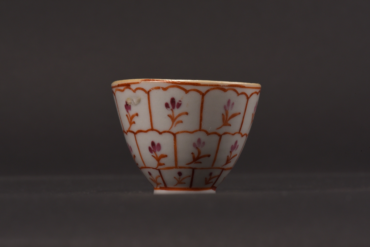 Kopp av vitt dekorerat porslin.
Ostindisk slät kopp med en låg oglaserad fotring.
Koppens utsida är dekorerad i två röda nyanser, med ett rutmönster och stiliserade blommor.