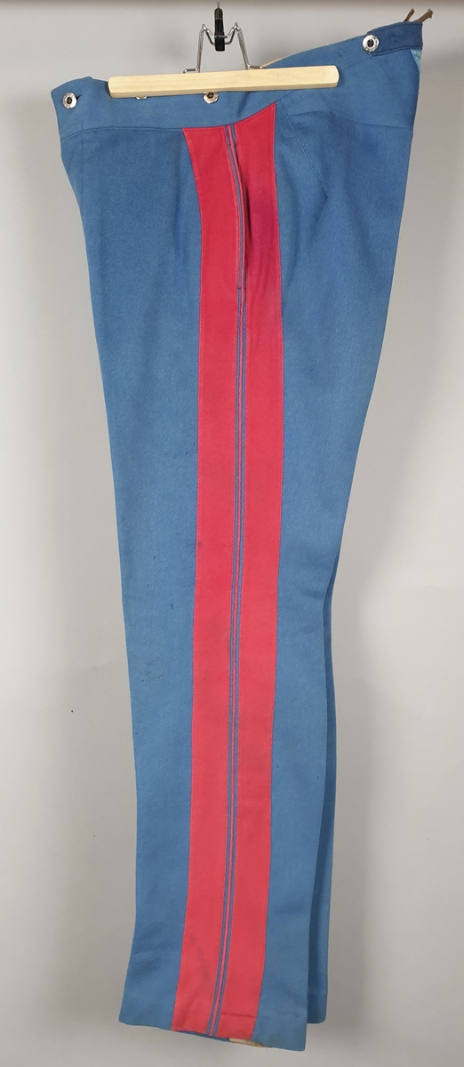 Blå uniformsbukse med røde striper på yttersiden av beina. Knapper i gylfen og innsydde lommer i sidene.